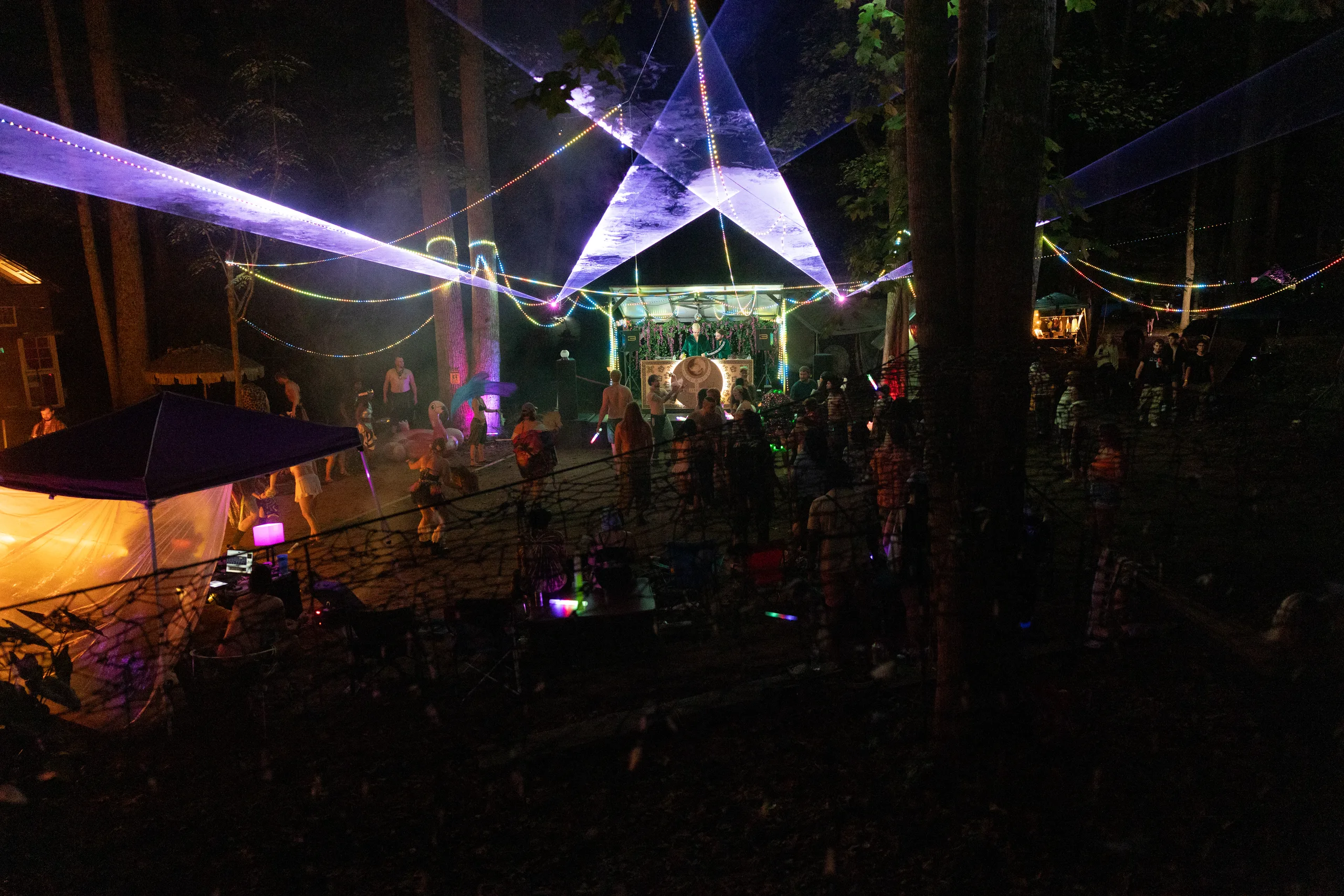 A forest dance floor beneath sweeping laser beams and string lights at ManaFest 2026