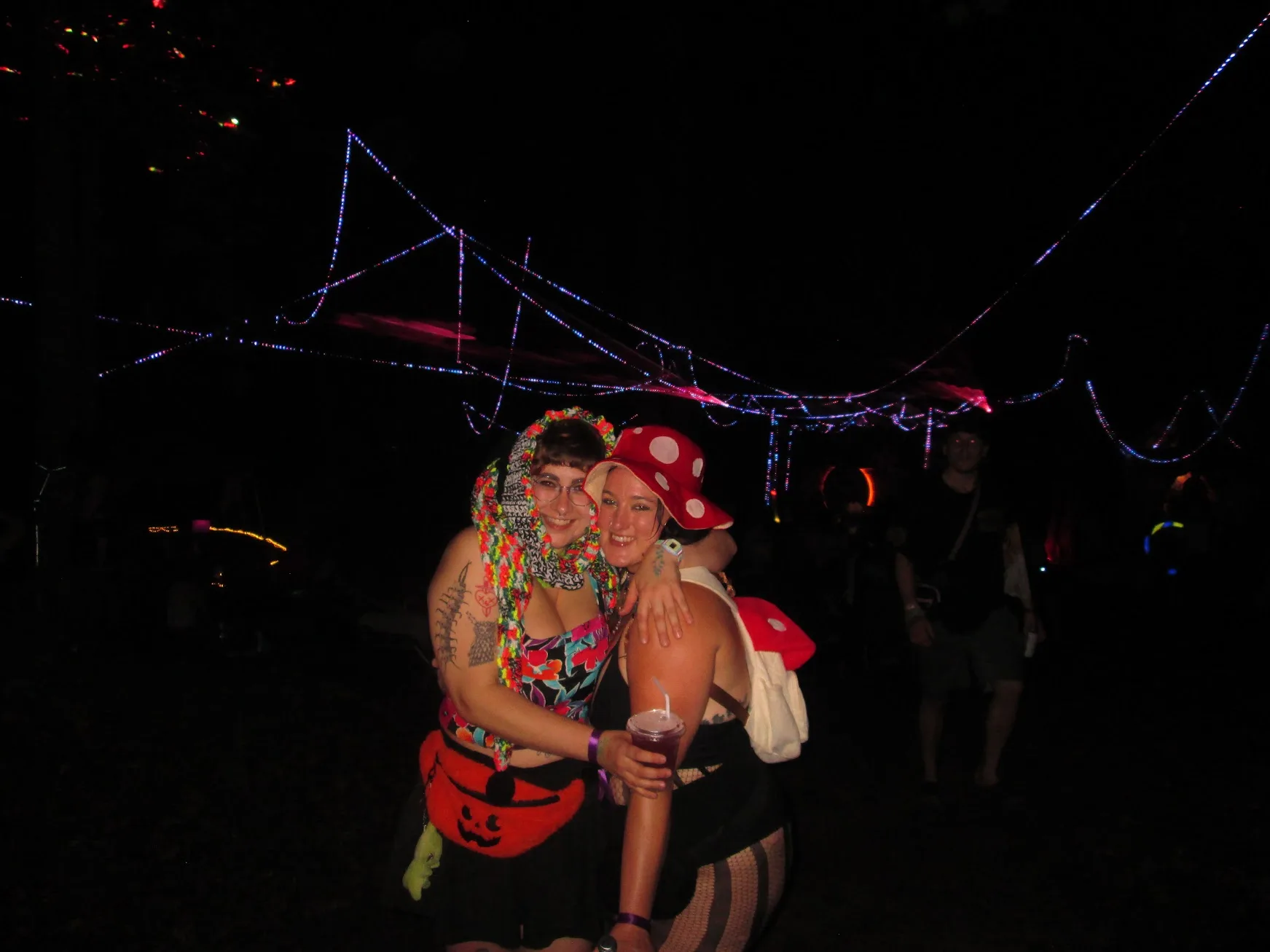 Two friends smiling beneath colorful string lights, one wearing a mushroom hat